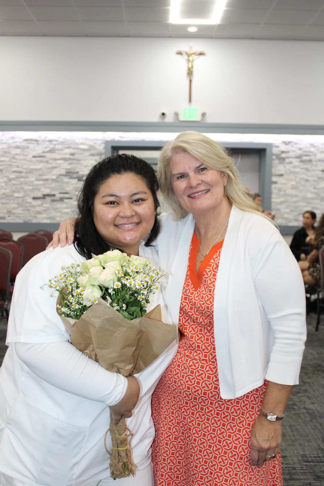A woman in white holds a bouquet while posing with another woman at a ceremony.