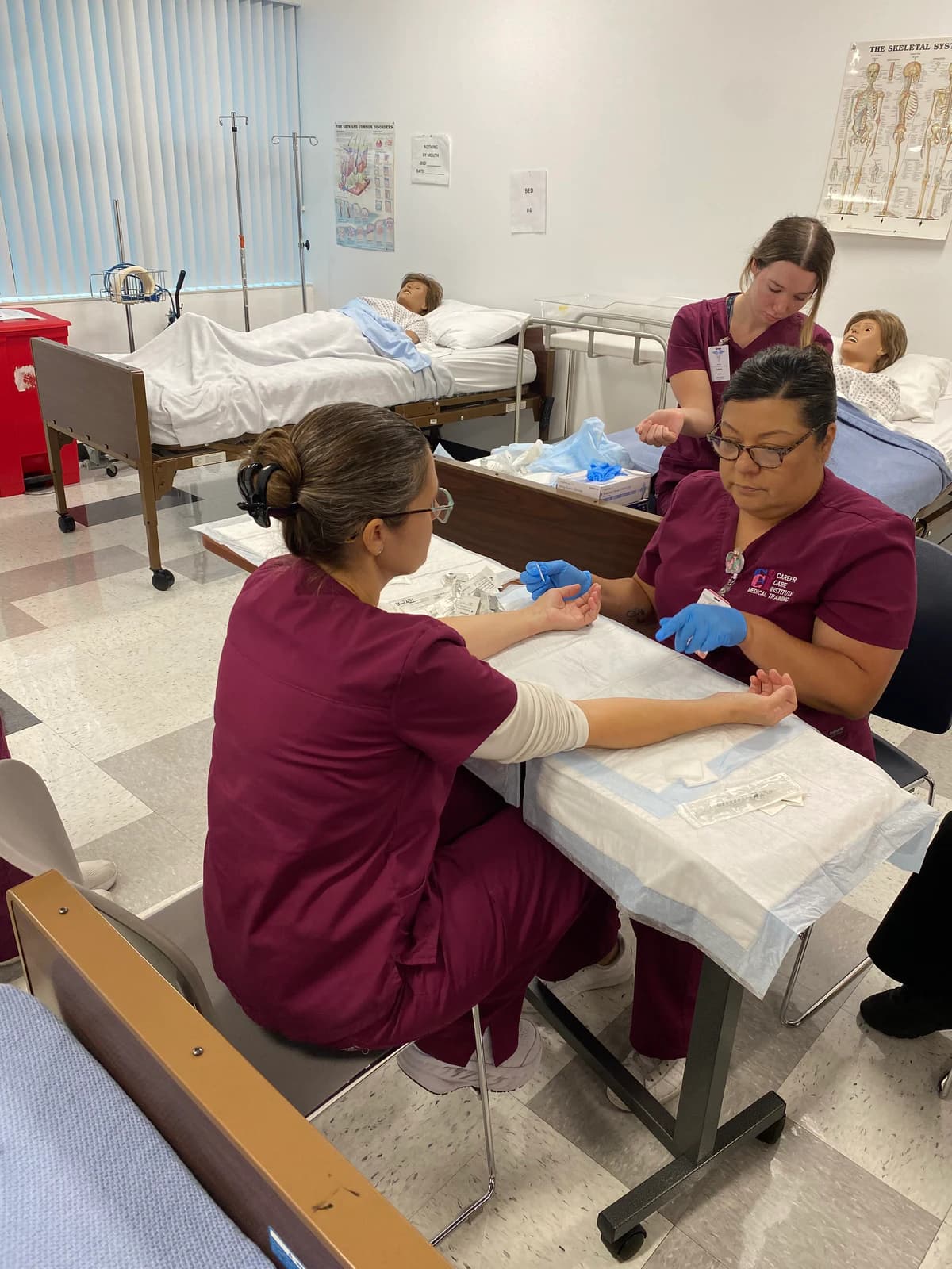 Vocational Nursing students in burgundy scrubs practice injection skills together in a nursing lab.