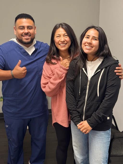Three people pose together indoors, one wearing blue scrubs.
